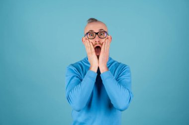 Balıkçı yakalı, kırmızı sakallı ve mavi arka planda gözlüklü genç bir adam kameraya bakar, ağzı açık şaşırır.