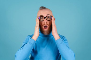 Balıkçı yakalı, kırmızı sakallı ve mavi arka planda gözlüklü genç bir adam kameraya bakar, ağzı açık şaşırır.