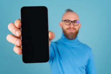 Mavi arka planda, boğazlı kazaklı, kırmızı sakallı genç bir adam odak noktasında bir yazı için boş siyah ekranlı bir telefon tutuyor ve kameraya uzanıyor.