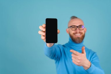 Mavi arka planda, boğazlı kazaklı, kırmızı sakallı genç bir adam odak noktasında bir yazı için boş siyah ekranlı bir telefon tutuyor ve kameraya uzanıyor.