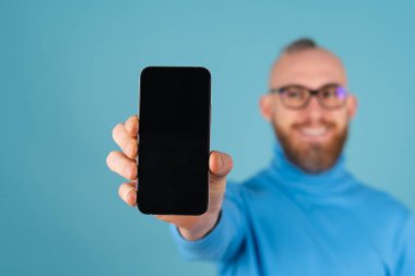 Mavi arka planda, boğazlı kazaklı, kırmızı sakallı genç bir adam odak noktasında bir yazı için boş siyah ekranlı bir telefon tutuyor ve kameraya uzanıyor.