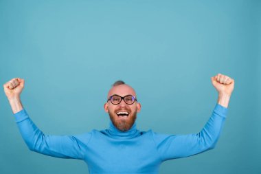 Mavi arka planda boğazlı kazaklı, kırmızı sakallı genç bir adam neşelidir, mutluluktan haykırır, kazanan bir jesttir.