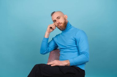 Mavi arka planda boğazlı, kırmızı sakallı genç bir adam ciddi ve katı bir yüzle bir sandalyede oturuyor.