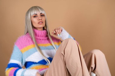 Uzun saçlı, kaküllü, çıplak makyajlı ve bej arka planda çok renkli, pofuduk bir süveter giyen bir sarışın neşeli bir şekilde gülümsüyor.