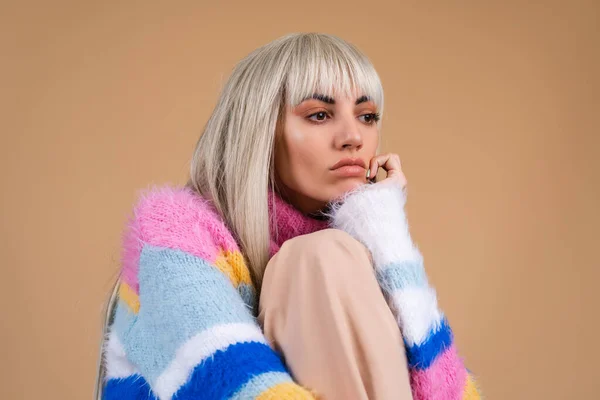 A blonde with long hair and bangs, with nude makeup and in a multi-colored bright fluffy sweater on a beige background, sad looks nowhere