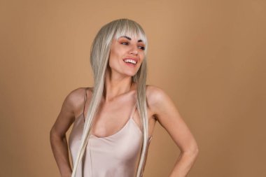 Sarışın, uzun saçlı, kaküllü, çıplak makyajlı ve bej arka planda ipek bir elbisesi olan, güler yüzlü, mutlu.