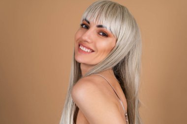 Sarışın, uzun saçlı, kaküllü, çıplak makyajlı ve bej arka planda ipek bir elbisesi olan, güler yüzlü, mutlu.