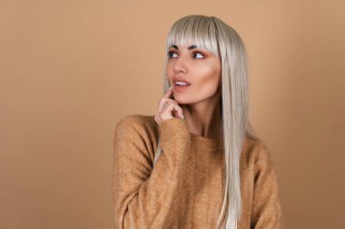 A blonde girl with bangs and brown daytime make-up in a sweater on a beige background looks thoughtfully to the left clipart