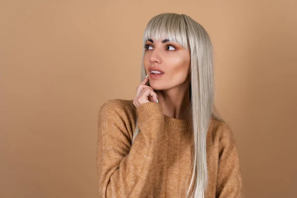 A blonde girl with bangs and brown daytime make-up in a sweater on a beige background looks thoughtfully to the left