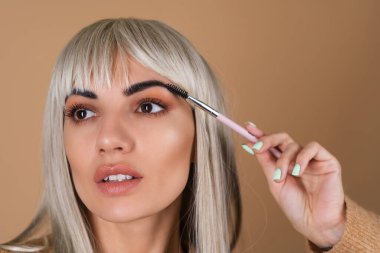 Kahverengi gündüz makyajlı ve kâküllü sarışın kız bej arka planda kaş fırçası tutuyor.