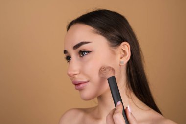 Bej bir arka planda genç bir kadının portresi. Doğal sıcak makyajlı ve pürüzsüz ciltli. Elinde allık için fırça tutuyor.