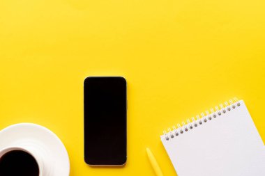 Üst kattaki ofis masasında yatıyordu. Not defterli çalışma alanı, ofis malzemeleri, kalem, cep telefonu boş ekran, sarı arka planda fotokopi alanı olan kahve fincanı.