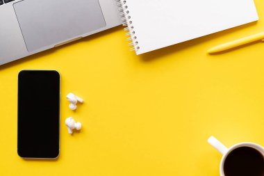 Üst kattaki ofis masasında yatıyordu. Not defteri, ofis malzemeleri, kalem, dizüstü bilgisayar, cep telefonu, sarı arkaplanda kopya alanı olan kablosuz kulaklık.