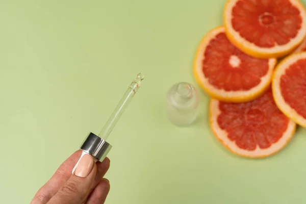 Natural vitamin C serum, skin care, essential oils. Cosmetic glass bottle with dropper and pieces of fresh juicy grapefruit. A woman\'s hand holds a pipette.