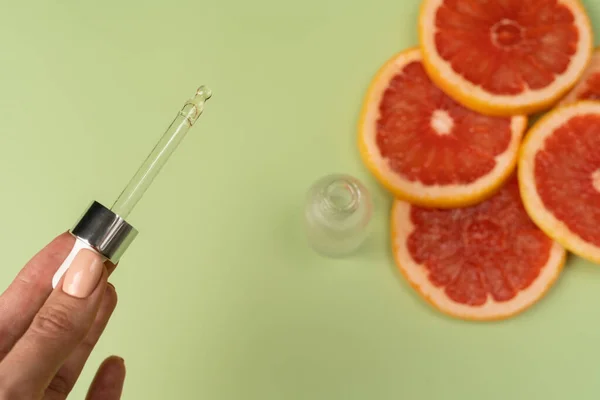 Natural vitamin C serum, skin care, essential oils. Cosmetic glass bottle with dropper and pieces of fresh juicy grapefruit. A woman\'s hand holds a pipette.