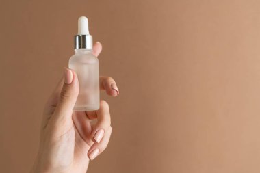 İnce parmakları ve modaya uygun pastel bej renginde kahverengi arka planda çıplak manikürü olan bir kızın zarif ellerinde bir yüz yağı serumu var.