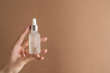 İnce parmakları ve modaya uygun pastel bej renginde kahverengi arka planda çıplak manikürü olan bir kızın zarif ellerinde bir yüz yağı serumu var.