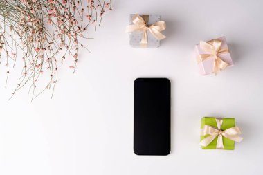 Düz yatış, üst manzara, beyaz arka plan, küçük hediye kutuları ve kurumuş çiçek çalıları 8 Mart için pembe sevimli çiçekler, bahar havası, boş siyah ekranlı telefon