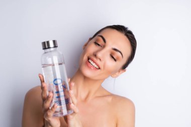 Mükemmel ciltli, doğal makyajlı, beyaz bir arka planda kişisel cam şişe su taşıyan üstsüz bir kadının güzel portresi.