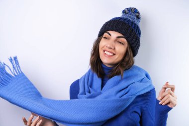 Mavi bir balıkçı yaka, şapka ve atkı giymiş beyaz bir arka planda neşeli bir kadın.