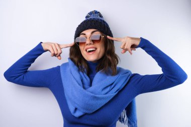 Mavi balıkçı yaka, şapka ve atkı takan, güneş gözlüklü, beyaz arka planda neşeli, neşeli bir kadın.