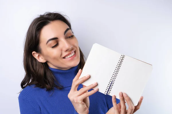 Young woman in a blue golf turtleneck on a white background pensive with a notebook in her hand thinks about ideas, goals for the year
