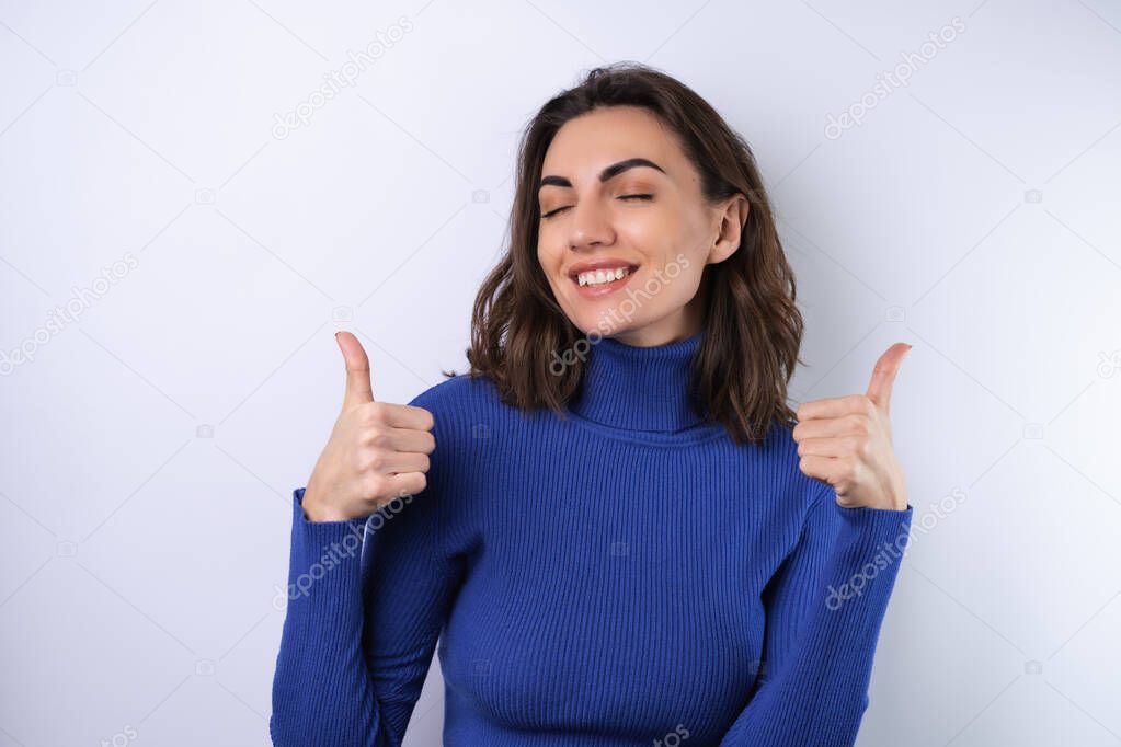Young woman in a blue golf turtleneck on a white background with a confident smile smiling cheerfully showing thumbs up