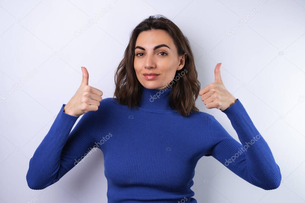 Young woman in a blue golf turtleneck on a white background with a confident smile smiling cheerfully showing thumbs up