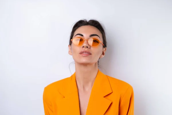 流行のスタイリッシュなメガネと白の背景に明るいオレンジの特大のジャケットの若い女性 — ストック写真