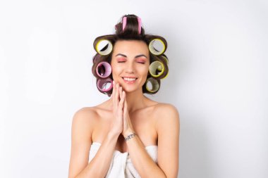 Güzel ev hanımı. Saç bigudileri ve çıplak omuzları olan neşeli genç bir kadın randevu gecesine hazırlanıyor. Beyaz bir arka planda evde saç stili yapar..