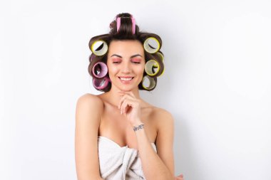 Güzel ev hanımı. Saç bigudileri ve çıplak omuzları olan neşeli genç bir kadın randevu gecesine hazırlanıyor. Beyaz bir arka planda evde saç stili yapar..