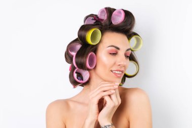 Güzel ev hanımı. Saç bigudileri ve çıplak omuzları olan neşeli genç bir kadın randevu gecesine hazırlanıyor. Beyaz bir arka planda evde saç stili yapar..