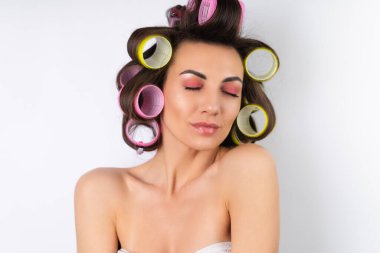 Güzel ev hanımı. Saç bigudileri ve çıplak omuzları olan neşeli genç bir kadın randevu gecesine hazırlanıyor. Beyaz bir arka planda evde saç stili yapar..