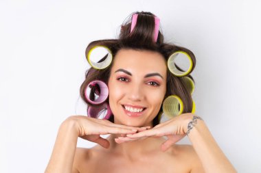 Güzel ev hanımı. Saç bigudileri ve çıplak omuzları olan neşeli genç bir kadın randevu gecesine hazırlanıyor. Beyaz bir arka planda evde saç stili yapar..