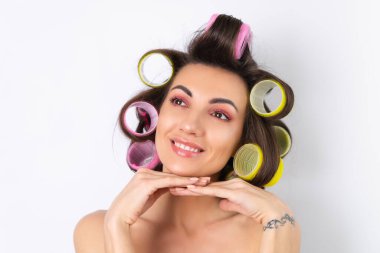 Güzel ev hanımı. Saç bigudileri ve çıplak omuzları olan neşeli genç bir kadın randevu gecesine hazırlanıyor. Beyaz bir arka planda evde saç stili yapar..