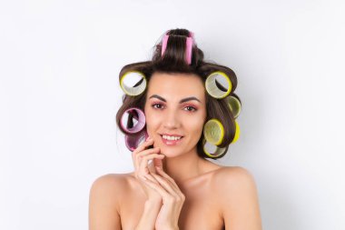 Güzel ev hanımı. Saç bigudileri ve çıplak omuzları olan neşeli genç bir kadın randevu gecesine hazırlanıyor. Beyaz bir arka planda evde saç stili yapar..