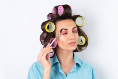 Güzel ev hanımı. Saç bigudileri ve kaş fırçası olan neşeli genç bir kadın randevu gecesine hazırlanıyor. Beyaz bir arka planda evde saç stili yapar..