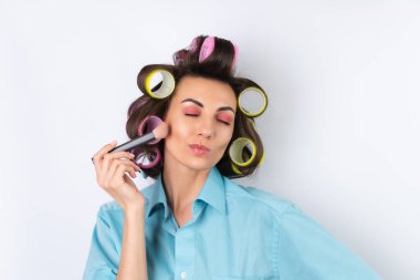 Güzel ev hanımı. Saç bigudileri, parlak pembe makyajı ve pamuklu fırçası olan neşeli genç bir kadın beyaz bir arka planda bir randevu gecesi, akşam yemeği için hazırlanıyor..