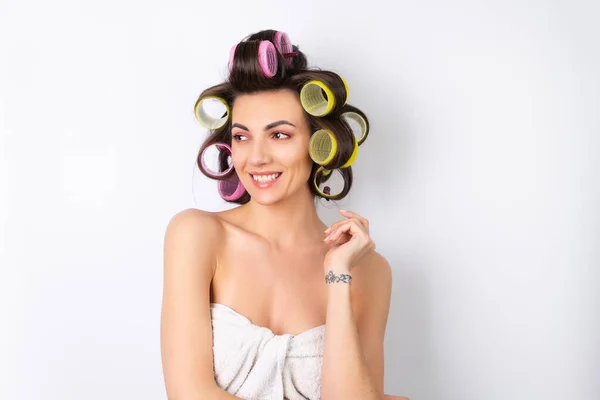 Beautiful housewife. Young cheerful woman with hair curlers and bare shoulders getting ready for a date night. Makes a hairstyle at home on a white background.