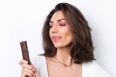 Kokteyl elbiseli, altın zincirli, beyaz arka planda parlak pembe makyajlı genç bir kadın. Sağlıklı bir mısır gevreği çikolatası tutar..