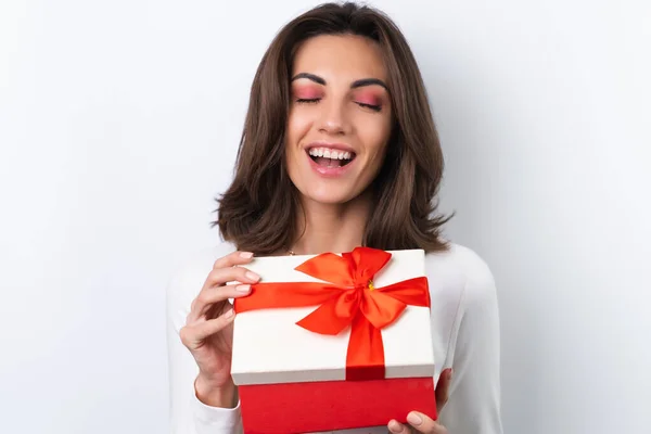 Young woman in a cocktail dress, gold chain, bright spring pink makeup on a white background. Holds a gift box for March 8 and smiles cheerfully.
