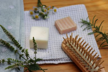 Sıfır atık kozmetik. Doğal organik katı el yapımı şampuan sabunlar ve nane, papatya çayı ve biberiye bitkisi havluların yanında ve ahşap masa üzerinde ahşap saç fırçası.