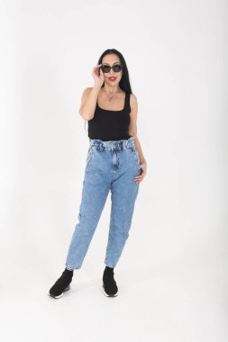 Stüdyoda güneş gözlüklü ve kot pantolonlu beyaz arka planda şık bir kadın.