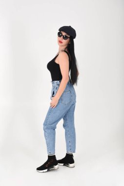 Stüdyoda beyaz arka planda, güneş gözlüklü, kot pantolonlu, siyah tişörtlü ve siyah şapkalı şık bir kadın var.