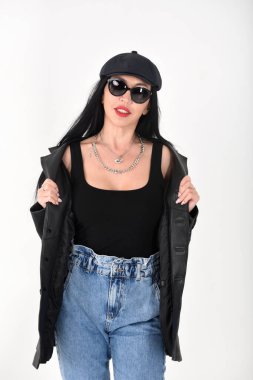 Beyaz arka planda, güneş gözlüğü, kot pantolon, siyah tişört, siyah şapka ve siyah deri ceket giyen şık bir kadın.