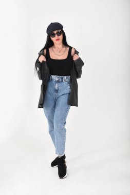 Beyaz arka planda, güneş gözlüğü, kot pantolon, siyah t-shirt, siyah şapka ve siyah deri ceket giyen şık bir kadın.