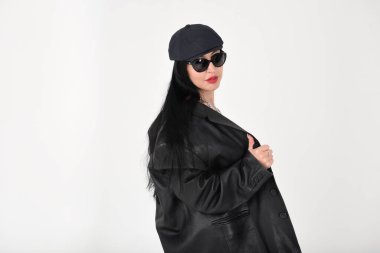 Stüdyoda beyaz arka planda, güneş gözlüğü takan, siyah şapkalı ve siyah deri ceketli şık bir kadın.