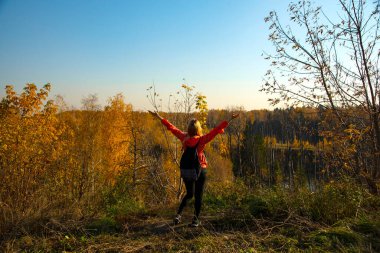 Sırt çantalı güzel bir sarışın, bir orman parkında, sarı sonbahar yapraklarının arka planında güzel bir gülümsemeyle yürüyor.