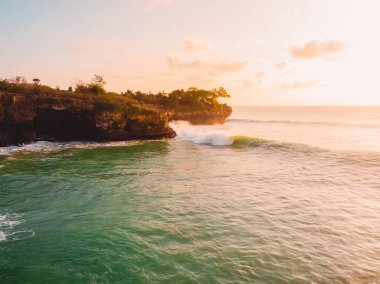 Sörf dalgaları ve sıcak gün batımıyla kayalık kıyı şeridinin hava manzarası.
