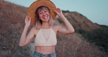 Hasır şapkalı, mutlu bir şekilde kameraya bakan gülümseyen kadının portresi gün batımında açık havada başarılı bir yaşam tarzının keyfini çıkarıyor.